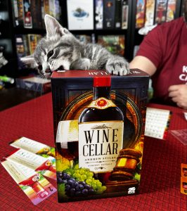 Game box for Wine Cellar standing up on a table with a gray kitten behind it trying to bite the upper right hand corner. 